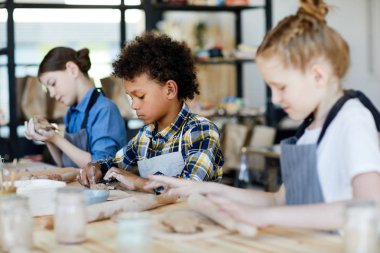 Mixed-race schoolboy sitting by table between his classmates and making something from clay clipart