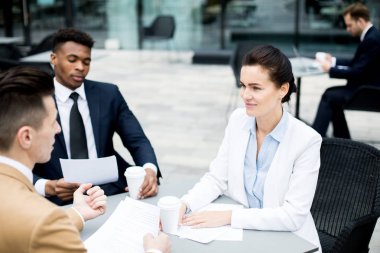 Çalışma noktalarının görüşülmesi için açık havada masa kenarında toplanan kağıt ve içeceklerile genç yöneticiler grubu