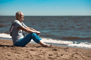 Yaşlı yalınayak adam kum üzerinde oturan ve deniz kenarında yaz tatili resort adlı arıyorum.