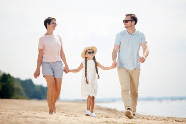 Yaz tatili sırasında sahilde yürüyüş keyfi yaparken sevimli küçük kız ile el ele mutlu aile tam uzunlukta portre