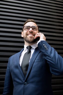 Genç, başarılı, iş elbisesi içinde gülümseyen, gözlüklü, konuşan cep telefonlu, siyah profilli çarşafların yanında duran ve yüzünü çeviren yakışıklı bir erkek.