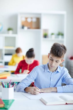 Mavi gömlekli çalışkan öğrenci, arka planda iki kızla birlikte not lar yapmak veya masa başında çizim yapmak