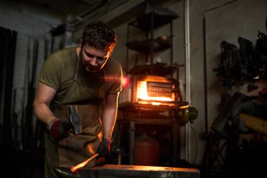 Demirci atölyesindeki genç adam atölyedeki örse yaslanırken demir dövmesi yapıyor.