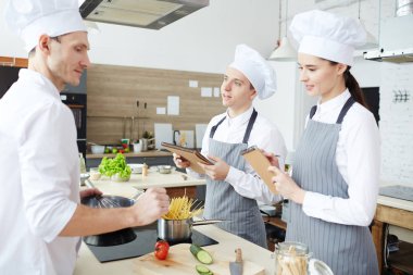 Restoran mutfağında menü oluşturan pozitif genç aşçılar grubu, önlükgenç çalışanlar yeni yemekler hakkında şef soran ve notlar yapma