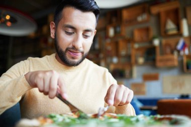 Young man licking his lips while cutting tasty pizza on plate by lunch in cafe