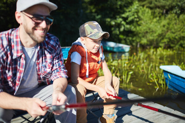 Удовлетворенный мальчик сидит на рыбалке вместе со своим отцом в солнечный летний день
