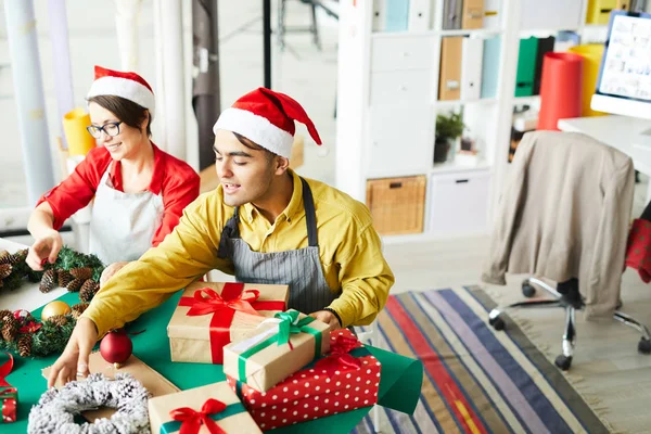 サンタキャップとエプロンの若い男と彼の同僚は クリスマスの装飾や梱包の贈り物を作る — ストック写真