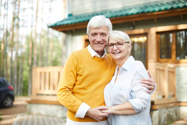 Forest house of grandparents: cheerful excited senior couple embracing each other and laughing while looking at camera and standing against cottage