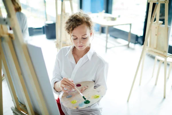 スタジオでアートクラス中にイーゼルに絵を描く前にパレットから濃い緑の色を取る若い女性 — ストック写真