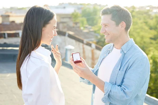 快乐的兴奋的年轻人把订婚戒指送给心爱的女人 要求她做他的妻子 微笑着的女孩挥手致意 不相信自己的眼睛 他们站在屋顶上 — 图库照片