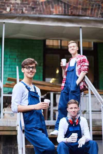 青いオーバーオールを着た若い工場労働者がポーチで休んで休憩時間にコーヒーを飲むと 彼らはカメラを見ている — ストック写真