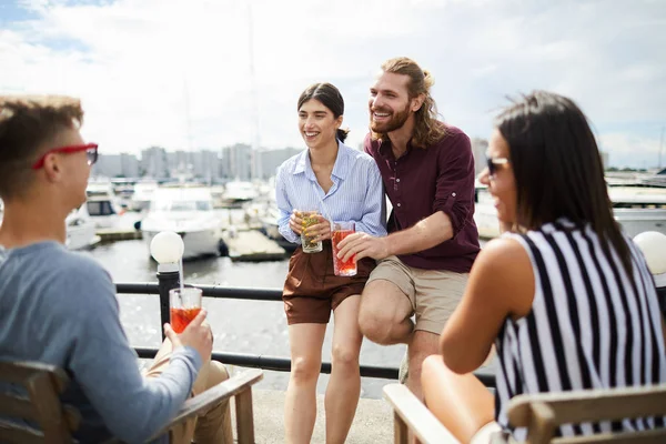 Happy young man and woman with drinks having talk to their friends by waterside on summer day