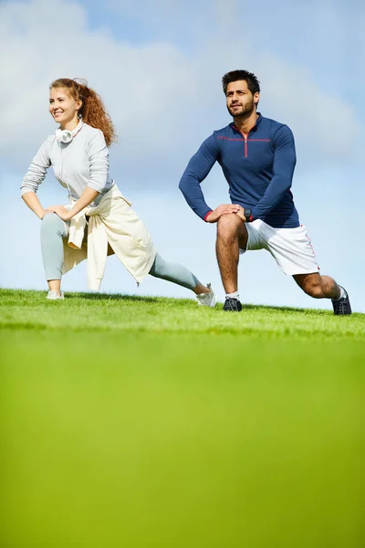 午前中に緑のフィールド上の脚のための運動をしている健康なアクティブ若いカップル — ストック写真