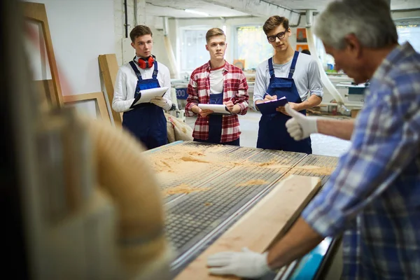 若い学生に森の働き方を説明しながら親指を立てた手袋をしたプロの先輩大工が ワークショップでゴムマットに置かれた板の表面をチェック — ストック写真