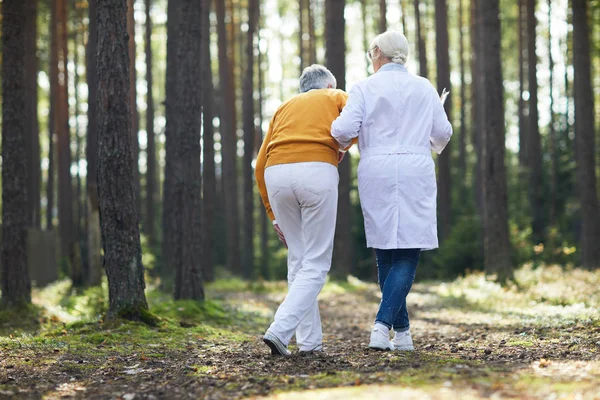 穿白大衣的女临床医生帮助病人走路 同时两人沿着森林小路移动 — 图库照片