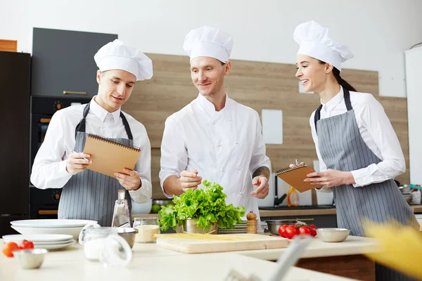 白い制服を着たコンテンツプロのシェフが料理教室で講義を行い 食べ物を持ってキッチンカウンターに立っている間に学生のメモをチェック — ストック写真