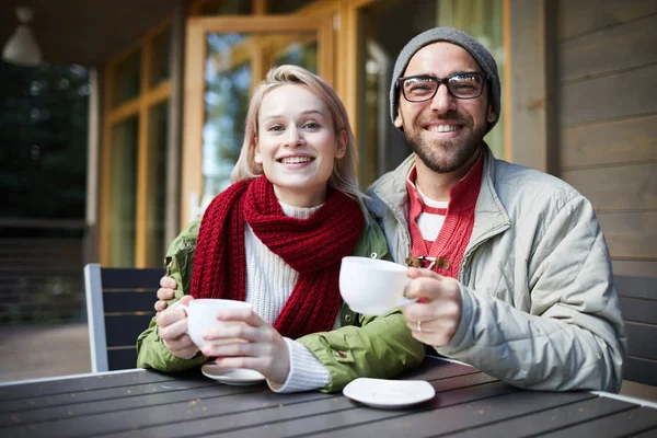 年轻美丽的白种人夫妇坐在门廊上的小桌子上 拿着一杯新鲜的咖啡 在镜头前高兴地微笑着 — 图库照片
