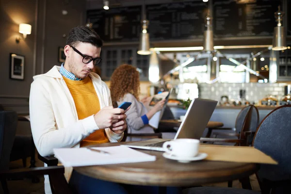 Casual Młody Biznesmen Przewijanie Smartfonie Siedząc Kawiarni Przed Laptopem — Zdjęcie stockowe
