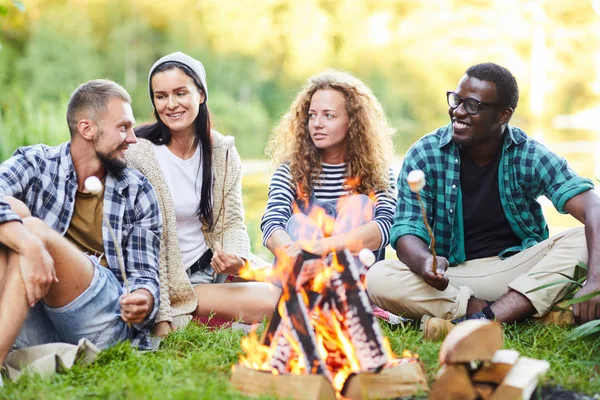 Group of young campers sitting by campfire and frying marshmellows while talking to each other