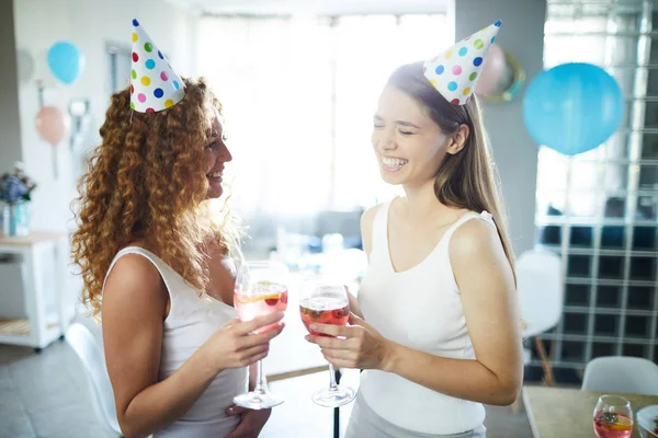 Carefree laughing girls in birthday caps and white clothes having homemade drinks at party