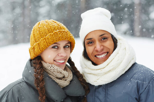 Smiling pretty multi-ethnic girls in stylish hats looking at camera and standing under snow outdoors