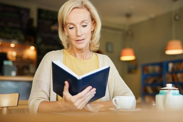 成熟的金发女性阅读书或她的笔记在记事本 而坐在咖啡馆 — 图库照片