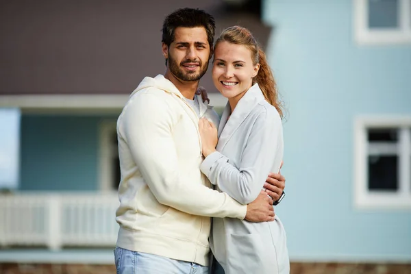 新しい家の背景にあなたを見ながら抱きしめる若い夫と妻 — ストック写真