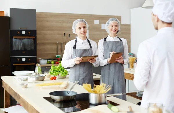 Cheerful cooking students in sterile caps and aprons standing against chef and listening to him while making notes during master class