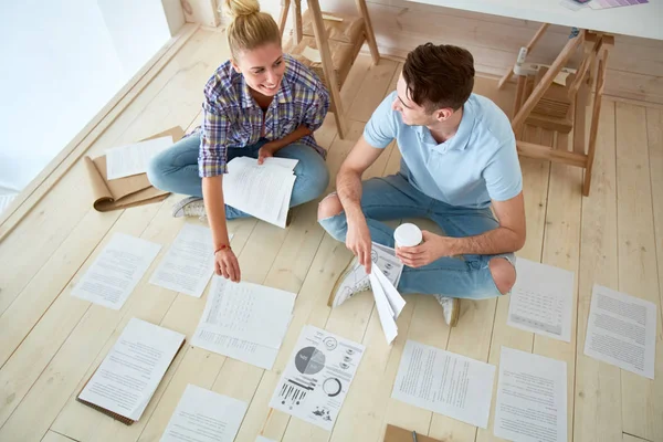 两名穿着休闲装的年轻员工坐在现代工作室的木地板上 用纸和谈话 — 图库照片