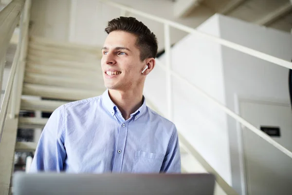 Young contemporary businessman in earphones sitting on staircase and enjoying music during work