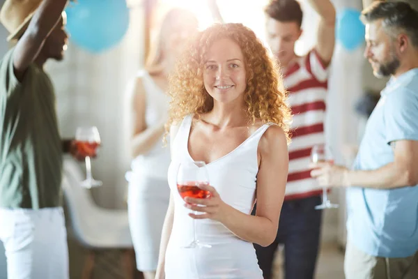 白いドレスを着た可愛い若い女性が ダンスの友人を背景に飲み物で応援しながらあなたを見ている — ストック写真