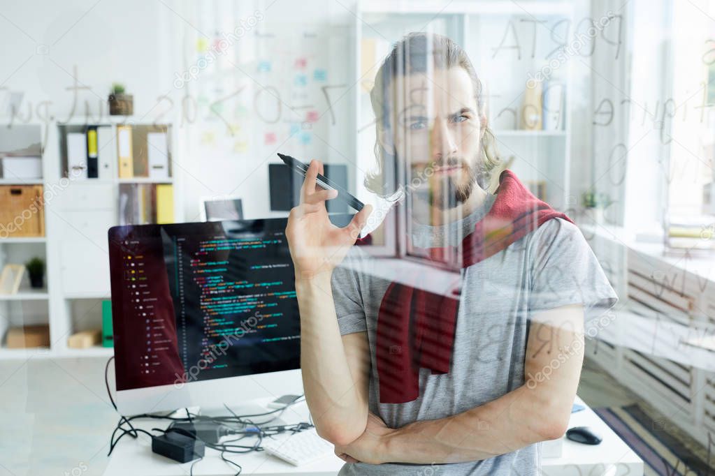 Serious confident handsome young computer programmer with beard developing code for software and holding felt tip pen while looking at data in glassy board in office