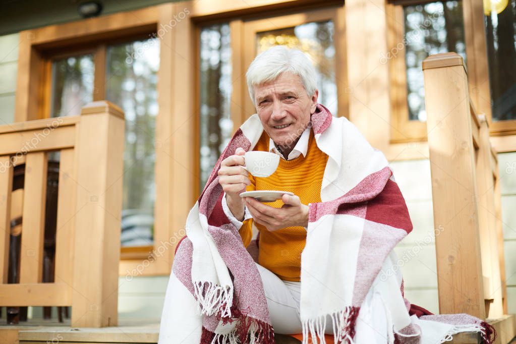 Content handsome gray-haired senior man covered with blanket sitting on porch of country house and drinking hot coffee to warm up