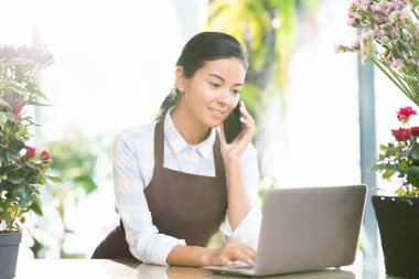 Dizüstü bilgisayar önünde online bilgi arayan ve istemciye telefonda konuşurken önlük içinde genç esmer kadın