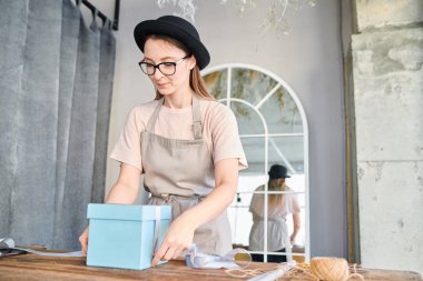 Genç kadın iş giysisi ve ahşap masa tarafından ayakta ve ipek kurdele ile mavi giftbox ipe şapka
