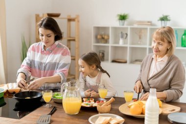 Kızı ve annesi için kahvaltı pişirirken sıcak tava üzerinde yumurta kırma genç kadın