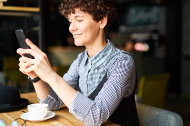 Smartphone içinde kaydırma ya da kafede çay yaparken video izlerken gündelik giysiler içindeki genç gülümseyen kadın