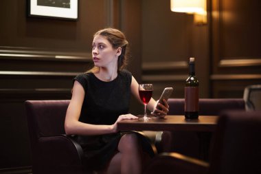 Girl in black dress looking aside while scrolling in smartphone and having red wine by table in classy restaurant clipart