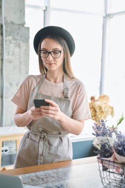 Genç ve güzel kadın önlük ve şapka smartphone içinde Studio müşteri siparişleri ile çalışırken ileti