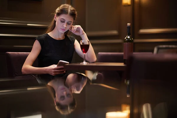 年轻优雅的女人与智能手机发短信和有葡萄酒 而坐在餐桌上在餐厅 — 图库照片