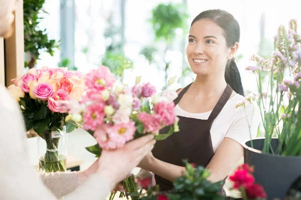 年轻的微笑的女人在工作服出售花束新鲜的粉红色玫瑰给她的客户在花店 — 图库照片