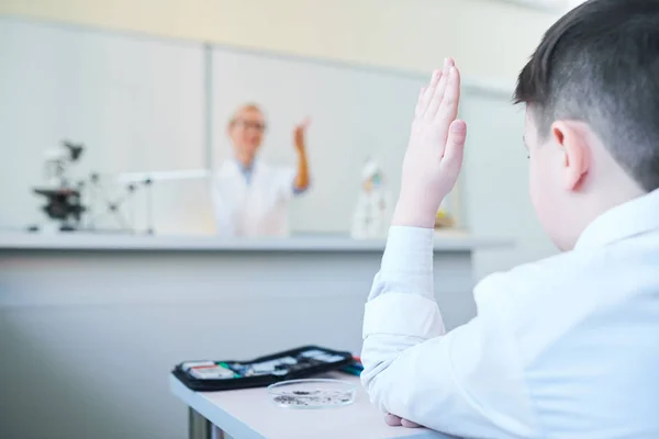 化学の授業で先生の質問に答えるために彼の手を上げる机によって若い学童 — ストック写真