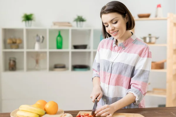 キッチンで野菜サラダのための新鮮なトマトをカットカジュアルなシャツで幸せな若い女性 — ストック写真