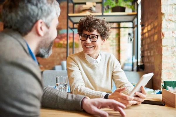 Cheerful young elegant businesswoman with tablet looking at co-worker while showing and explaining him online data