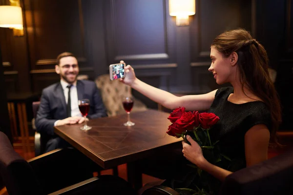 漂亮的女孩与一束红玫瑰做自拍 而坐在桌旁的男朋友在餐馆 — 图库照片