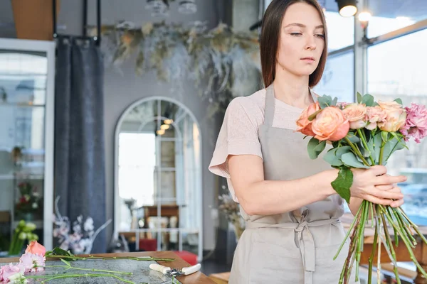 Young pretty female in workwear going to pack bunch of fresh roses while preparing flowers for sale