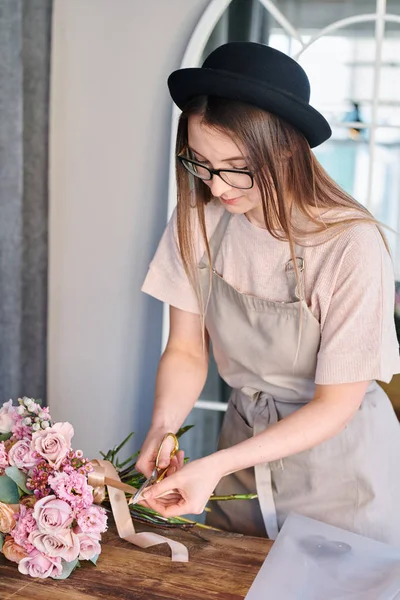 Pretty young florist cutting beige silk ribbon ends with scissors while making bouquet of pastel pink flowers in studio