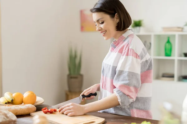 野菜サラダを準備しながらサラダ用のトマトをカットするカジュアルシャツの若い陽気な主婦 — ストック写真