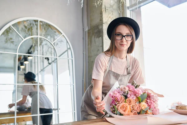 年轻的花商在眼镜 帽子和围裙看着你 同时包装一堆新鲜的柔和玫瑰 — 图库照片