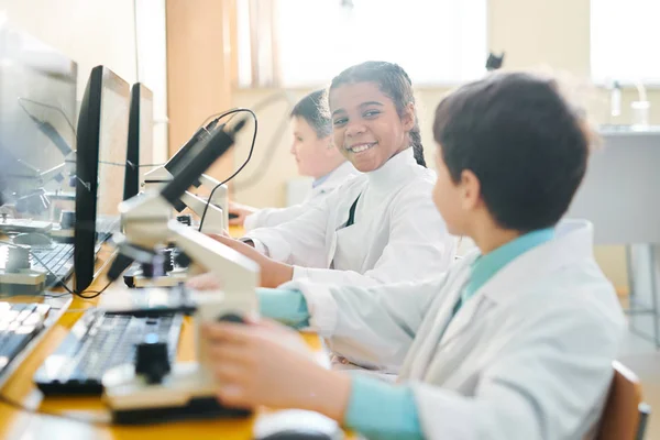 顕微鏡で作業しながら 化学のレッスンで彼女のクラスメートを見て幸せな小さな女子高生 — ストック写真